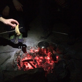 Campfire has reduced to coals - someone is pitting bread on a stick to toast while the billy boils
