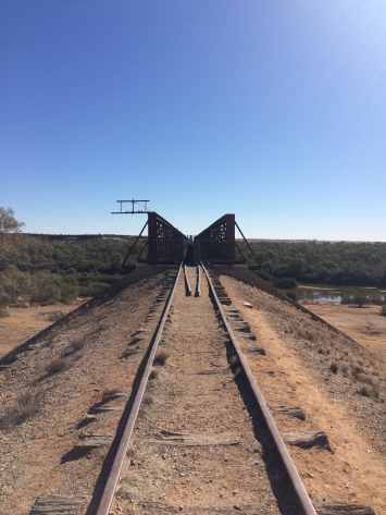 Algebuckina Bridge: photo taken standing on the disused railway tracks with the tall-sided bridge raised in front of green scrub etc on creek's far side, the flowing creek is also visible to the right. Sky is blue and everywhere else is dry clay-dirt