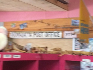 Blurred writing (thanks to camera focus) says "Oodnadatta Post Office", seen over the top of a pink shelf and on a pink wall.
