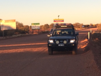 Camera looks back at two cars, the green-grey 4WD Nissan Pathfinder and maroon Nimbus Mitsubishi behind it, shadowed by the setting sun. Track behind them is brown-orange with signs to the left with info and warnings for travellers heading onto the Track