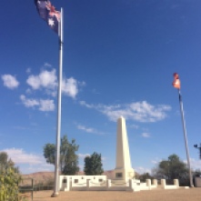 Flags on flagpoles stand next to a white stone obelisk reaching into the sky, with a plaque attached in memory of lives lost in war.