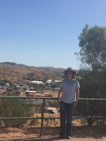 I'm standing in front of a steel fence - two bars across. Behind me is Alice Springs town. I'm wearing a blue collared t-shirt and jeans with my "jillaroo" wide-brimmed hat. I'm standing next to a green shrub and the sky is clear blue behind me.