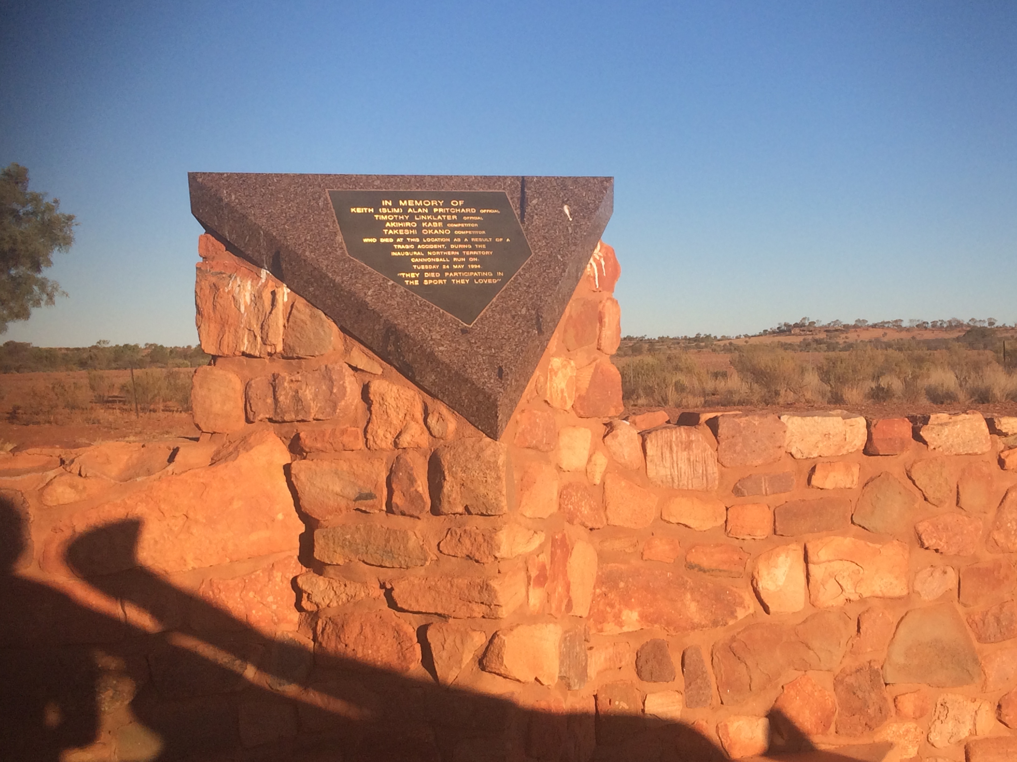 Red rock and mortar creating a fence with a raised corner which has a dark stone triangle on it. On the stone triangle is a plaque dedicated to those who lost their lives during the "Inaugural Cannonball Run"