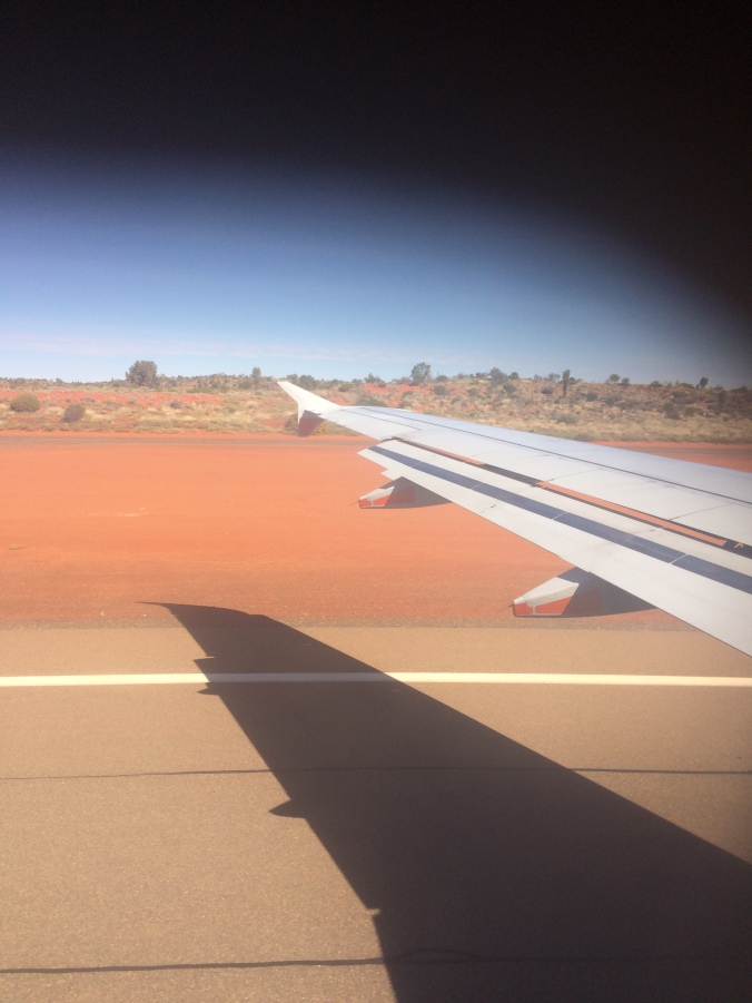 Red runway from the plane's window, also showing the wing and red dirt.