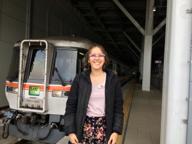 A woman (Clare) wearing glasses stands in front of a "wide-view" train. She is wearing a black coat over a pink top, with a colourful flowers-on-black-background skirt. Her hands are by her sides and she is smiling. The train is white with wide windows and a red stripe across its middle.