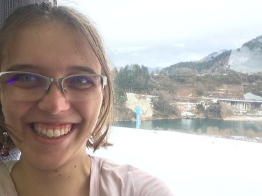 A woman (Clare) wearing glasses has taken a selfie smiling and the landscape shot shows the snow and lake with trees and mountain outside her window