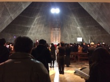 People, viewed from behind, are standing in a packed cathedral. The cathedral walls slowly slope up to a point at the top and a light shines above a cross.