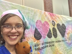 A woman (Clare)'s head and neck are visible to the left of the pic. A teddy (Womble)'s head rests next to Clare's neck. Clare is wearing a blue dress and pick-and-teal-tie-dye bandana. Behind her is a rainbow tie-dye fabric banner with words that read: MIV: 69th Intervarsity Choral Festival Melbourne 2018