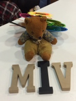 Womble, a brown bear with light brown pants and vest and blue shirt, sits on a white table supported by a rainbow flower-prop. In front of him sit wooden letters spelling, "MIV".