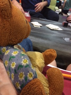 Womble, a brown bear with light brown pants and vest and blue shirt, sits on a red chair supported by a barely-visible human hand. In front of him is a black table with playing cards on it. Human hands and arms are visible on the opposite side of the table.