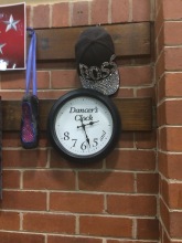 On a brick wall hangs a black ballet slipper on blue ribbon. Beside it are a black-and-silver cap with the word "Boss" written in silver on it, as well as a black and white clock that says, "Dancer's clock" in the middle, with "and 5 6 7 8" clockwise around its bottom edge.