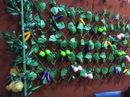 On a brick wall hangs a green and brown lattice frame with false green leaves attached. Tied to the frame are fake pieces of apple, eggplant, peach and carrot