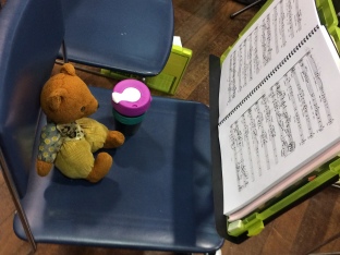 Viewed from side-on: Womble, a brown bear with light brown pants and vest and blue shirt, sits on a blue chair with a pink, teal and black KeepCup beside him. In front of him is an open music score on a music stand.