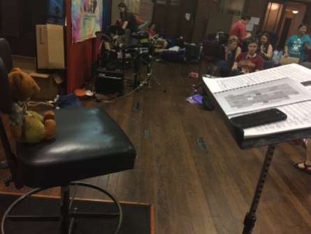 Viewed from side-on: Womble, a brown bear with light brown pants and vest and blue shirt, sits on a black stool. In front of him is a music stand with music score and, on top of that, a black phone and a booklet showing a black-and-white rehearsal schedule.