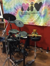 Womble, a brown bear with light brown pants and vest and blue shirt, sits on a black stool next to a drum kit. Two long drumsticks lay across his lap. Behind him is a red pinboard with a rainbow fabric banner on it. THe banner spells "MIV 69th Intervarsity Choral Festival Melbourne 2018"