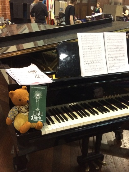 Womble sits on the edge of a black grand piano. The piano has a music score on its ledge and Womble is holding a green flyer advertising MIV's "Light the Dark" concert