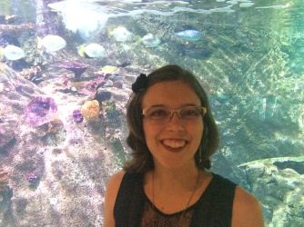 Clare's head and shoulders as seen as she stands in front of a coral fish tank. She is wearing a navy-blue dress, with her hair pulled back to the side with a black flower clip, and her glasses. The water behind her is very clear, with light reflected down through it shining onto the coral and fish.