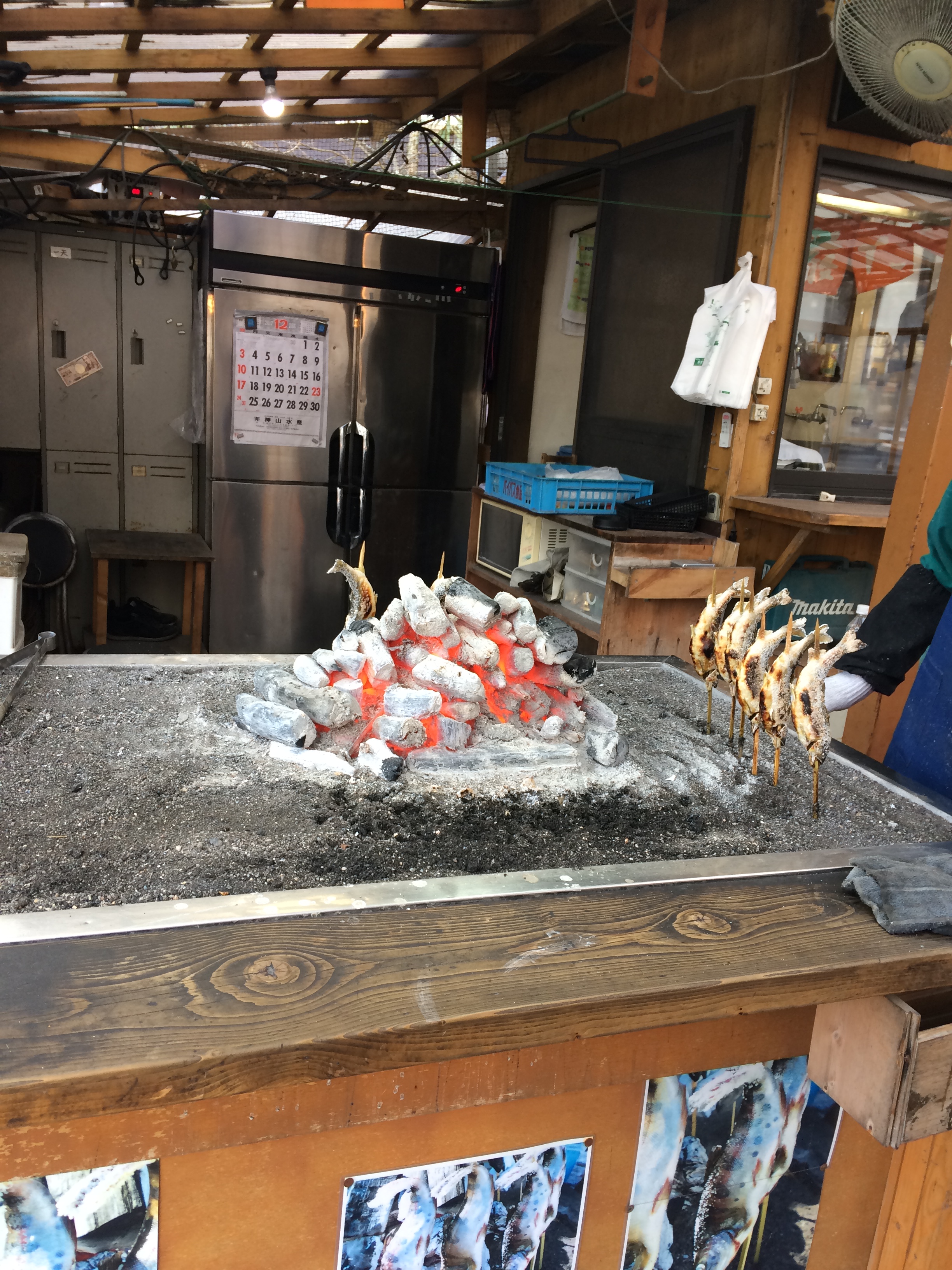 A heap of white coals sit atop a grate on a trolley. In the centre of the coals there is an orange colour from the heat. To the right side of and behind the pile of coals are rows of small whole fish, skewered through by sticks. A the back of the stall there are fridges visible and a flimsy roof supported b wooden beams is also seen.