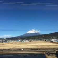 View from train - Mt Fuji's green slopes and snowy smoking peak in the background, a ploughed field in the foreground