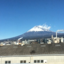Close-up view from train - Mt Fuji's green slopes and snowy smoking peak in the background rising above industrial suburbia
