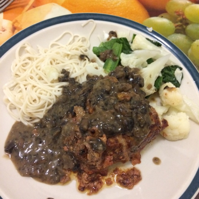 On a green-rimmed white plate sit a pile of white noodles, steamed cauliflower and Asian greens, along with meatloaf smothered in dark mushroom sauce