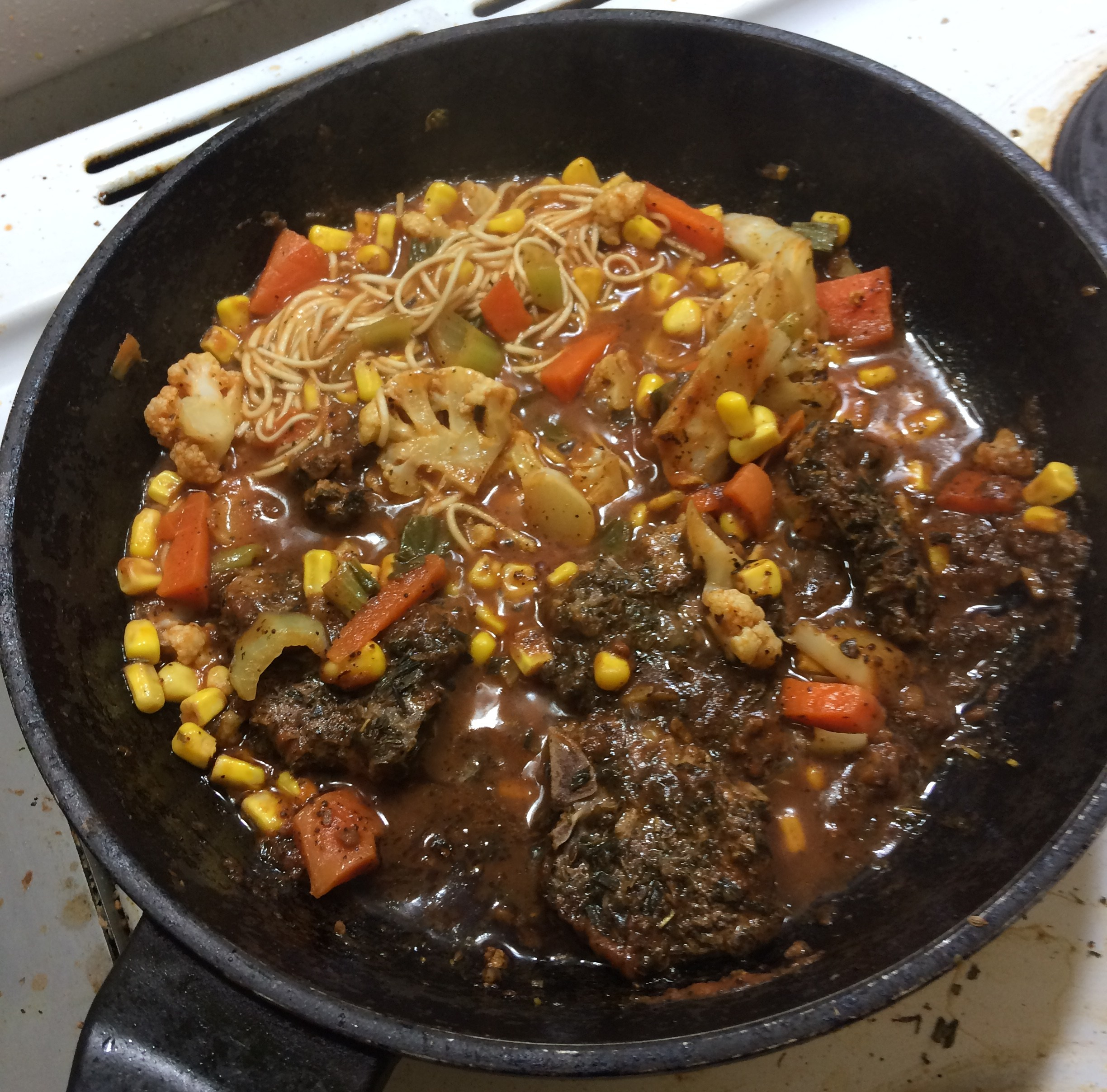 In a deep black frying pan are chops in a red tomatoey sauce with carrots, corn, bok choi and cauliflower with noodles stirred through