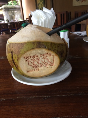 A whole coconut sits on a plate. Its husk has been removed and a hole cut in it to allow the insertion of a straw. On one side, a stylised design has been stamped on it - "bebel beng" with four duck silhouettes.