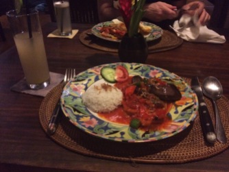 A glass of soft drink with a straw sit beside a plate with knife,, fork and spoon. On the colourfully-patterned plate are a ball of rice, chicken and vegetables in sauce.