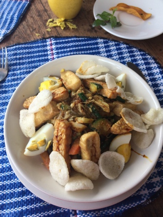 A white bowl sits on a blue striped tablecloth. In the bowl are eggs, tofu, tempeh and vegetables in a peanut sauce.